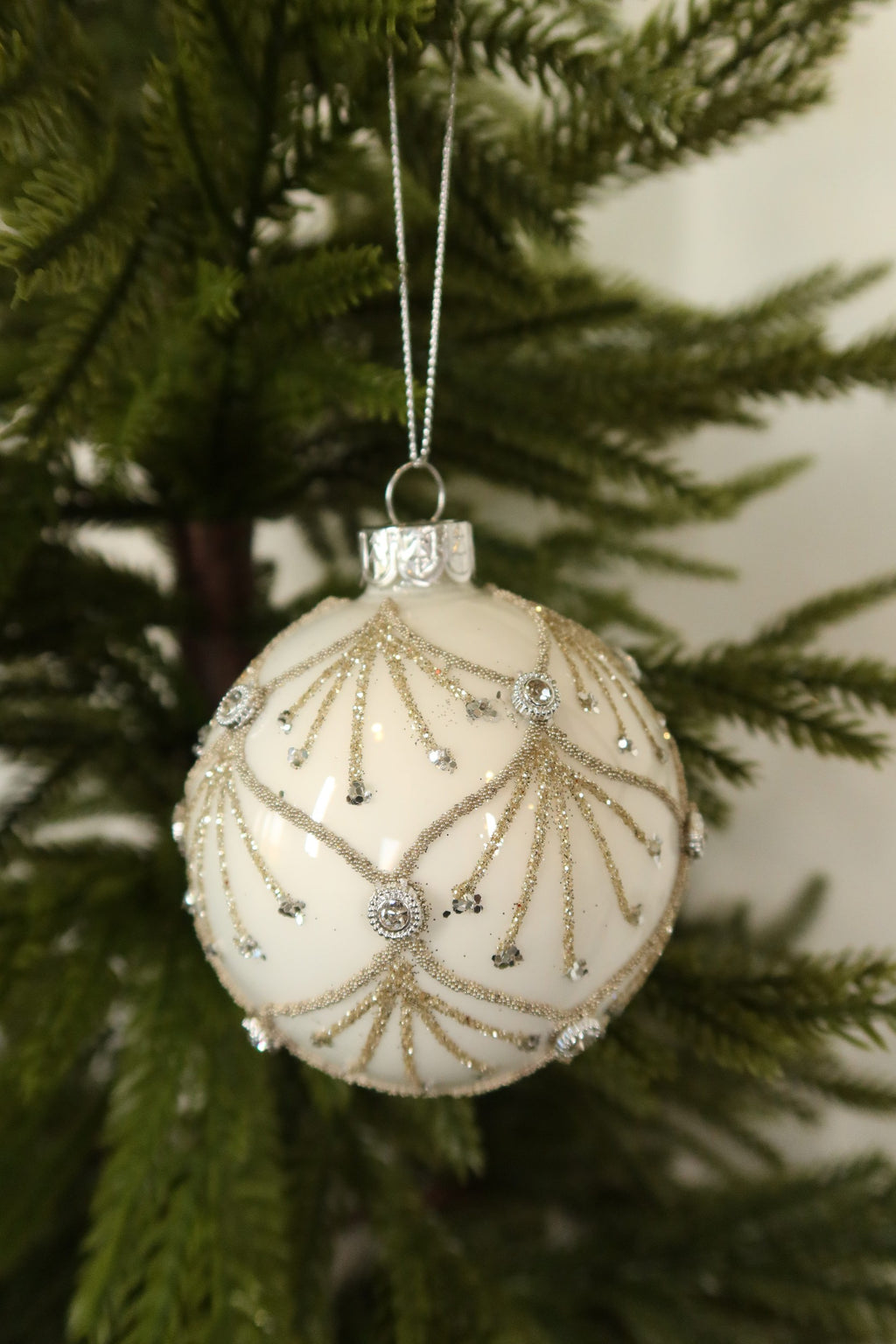 Front view of the white glass ball ornament with gold scallop design on a silver cord among green pine branches