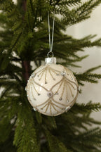 White glass ball Christmas ornament with gold glitter scallops and jeweled accents hanging on an evergreen tree
