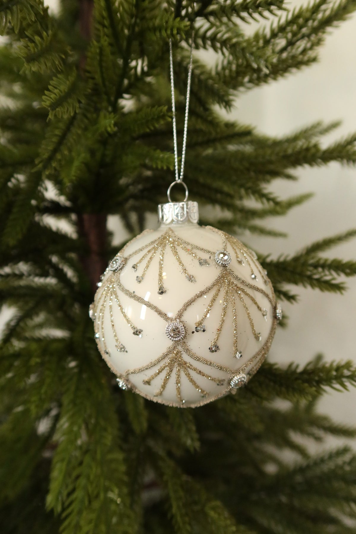 White glass ball Christmas ornament with gold glitter scallops and jeweled accents hanging on an evergreen tree
